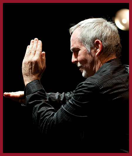Photo of a person in profile with one hand raised, appearing to conduct or gesture during a performance or presentation against a dark background.