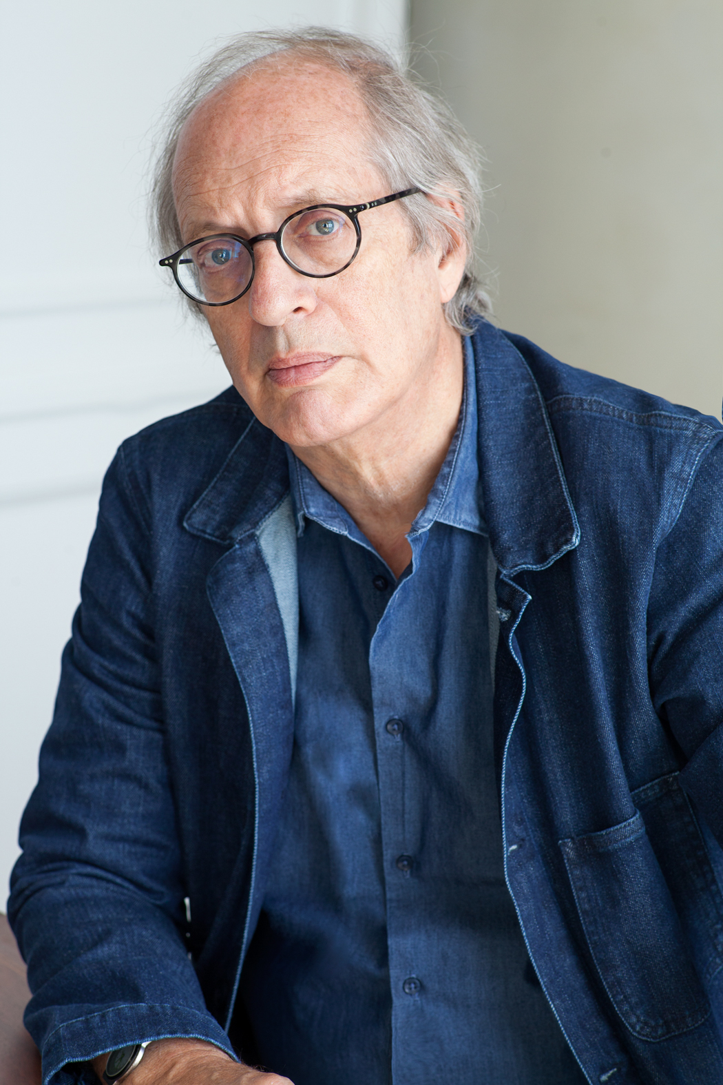 Portrait of a person with glasses wearing a blue shirt and jacket, seated indoors against a neutral background.
