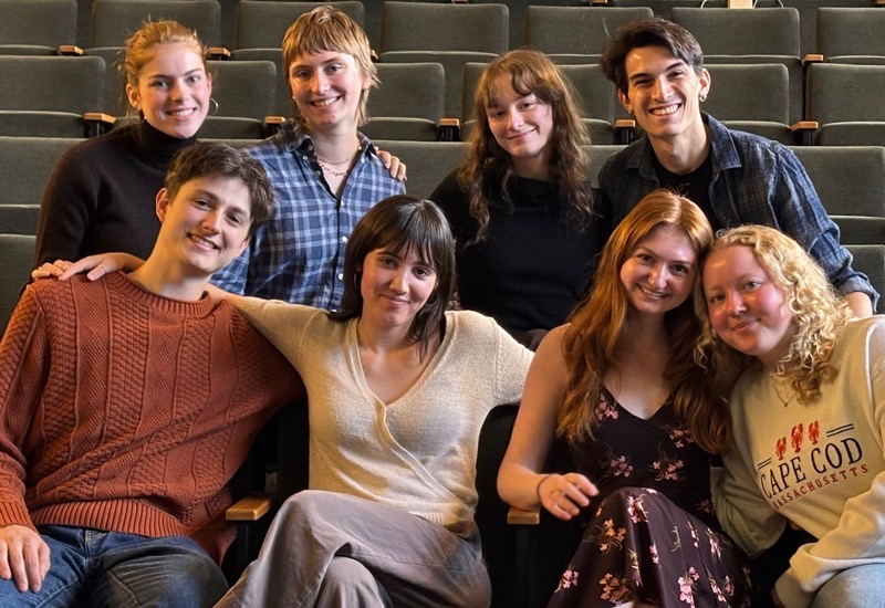 A group of eight individuals is seated in a theater setting. The foreground shows four seated figures, two on either side, with one figure extending an arm across the shoulders of another. The background features four standing figures, positioned in a semi-circle. The lighting is soft and warm, illuminating the faces and creating a relaxed atmosphere. The seating is arranged in a tiered fashion, enhancing the visual depth of the composition.
