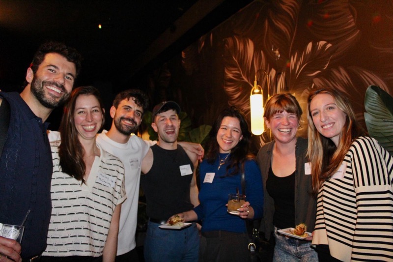 A group of seven individuals stands together in a dimly lit setting, displaying a variety of cheerful expressions. They are positioned closely, engaging in conversation while holding drinks and plates of food. The background features a large floral mural with rich, dark colors, and there are decorative light fixtures casting a soft glow. The overall atmosphere suggests a social gathering, enhanced by the lush greenery visible in the corners.