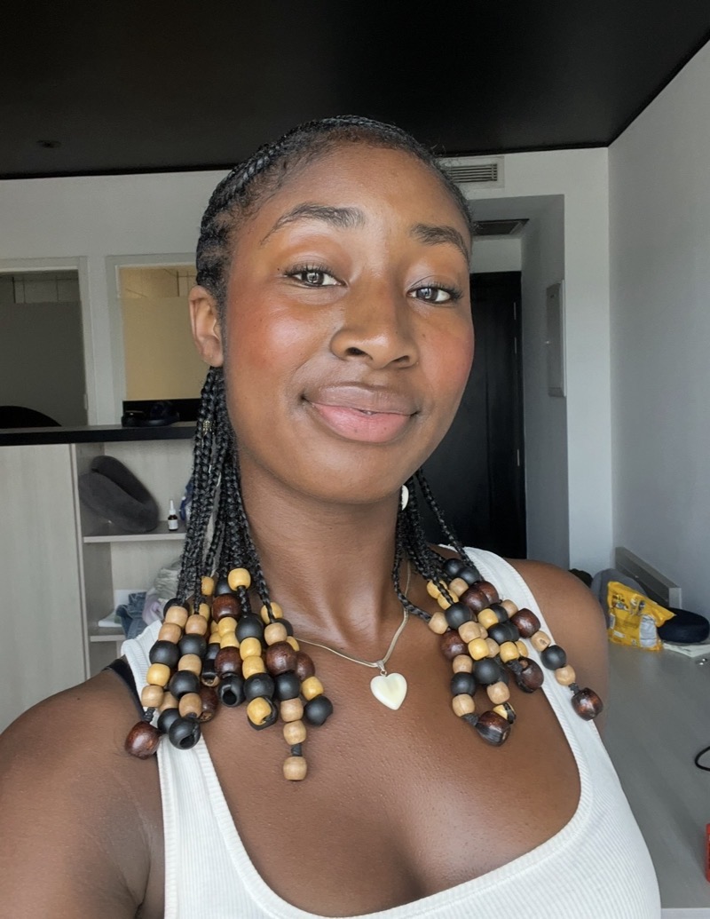 A close-up image features an individual smiling and looking directly at the camera, with braided hair adorned with colorful beads in hues of brown, yellow, and black. The individual wears a light-colored sleeveless top and a silver necklace with a heart-shaped pendant. The background includes light-colored walls and furniture, with soft lighting illuminating the scene. The overall composition captures the details of the hairstyle and the textures of the necklace and clothing against a subtle indoor setting.