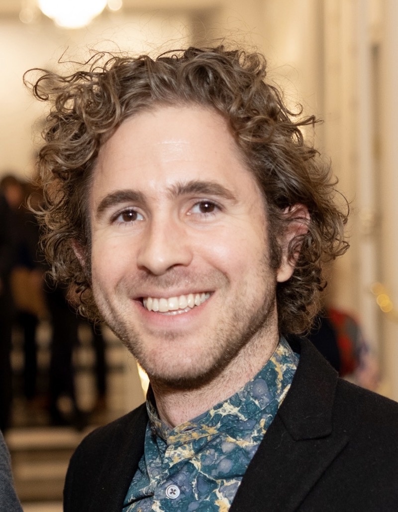 A person with curly hair smiles while wearing a patterned shirt featuring shades of blue and green. They are dressed in a dark blazer and are positioned in the foreground of a softly blurred interior space, where hints of warm lighting create a welcoming atmosphere. The background reveals indistinct figures and shapes, suggesting a lively gathering. The overall composition highlights the subject's expression and attire against a softly illuminated environment.