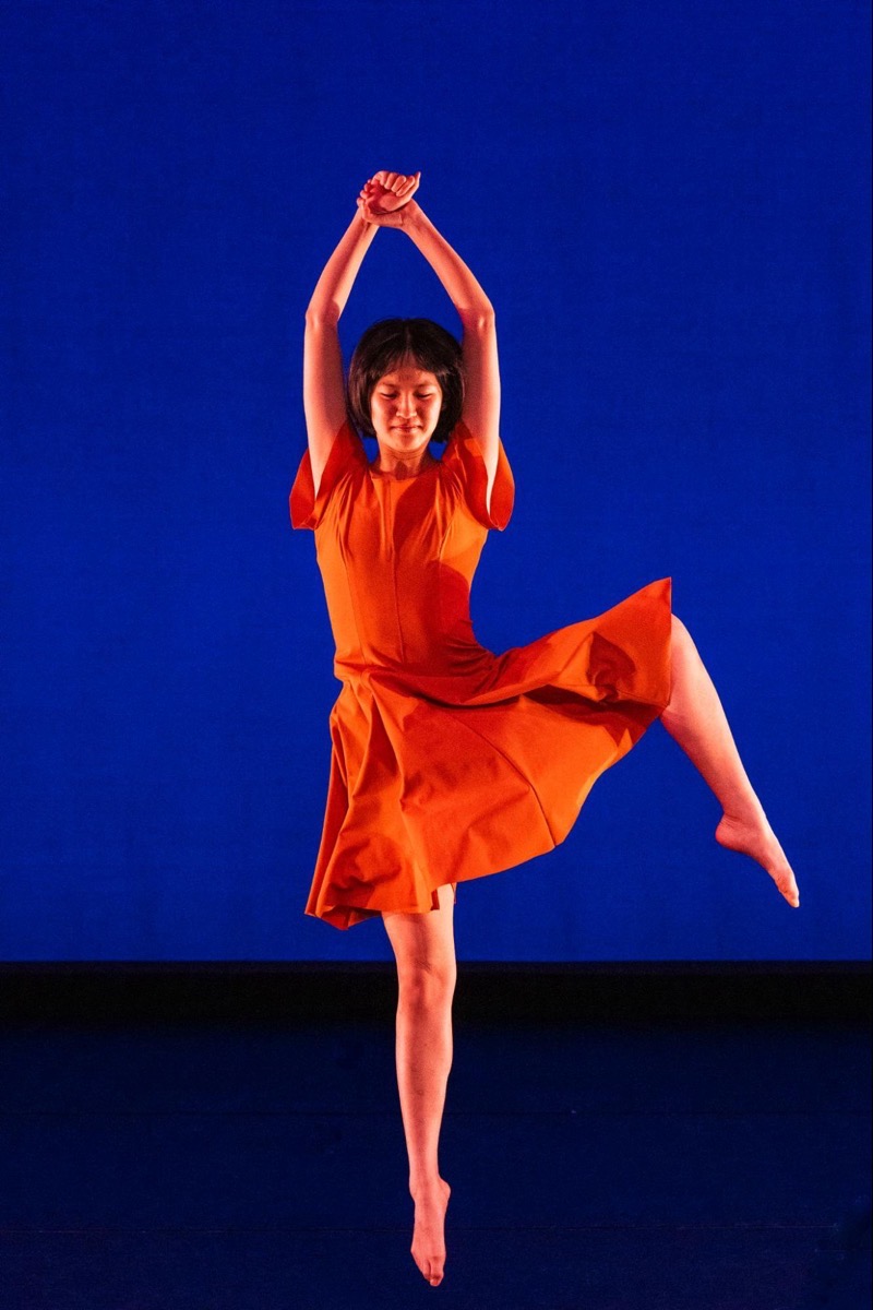 A figure in an orange dress gracefully poses against a vivid blue background. The arms are raised above the head, emphasizing a dynamic upward movement, while one leg is lifted, showcasing an elegant extension. The soft texture of the dress contrasts with the smoothness of the background, enhancing the visual interplay. Subtle lighting highlights the figure, creating depth and drawing attention to the fluidity of the pose.