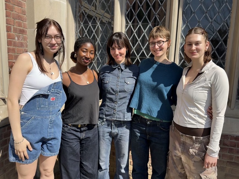 Five individuals stand side by side in front of a brick building adorned with large windows featuring diamond pane glass. They are positioned in casual attire: one wears denim overalls over a white top, another in a black tank, followed by a figure in a dark button-up shirt. The group is framed by sunlight, casting soft shadows on their clothing, and the warm tones of the brick contrast with the greenery visible through the windows, creating a vibrant, inviting atmosphere.