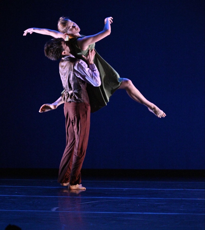 Two figures are engaged in a dance performance on a polished stage. One figure holds the other aloft in a dynamic pose, showcasing a balance of strength and grace. The figure being lifted wears a flowing green dress, while the other is dressed in a textured, lighter-colored garment. The background is a deep blue, enhancing the focus on the dancers and creating a dramatic contrast with the stage lit in soft highlights.