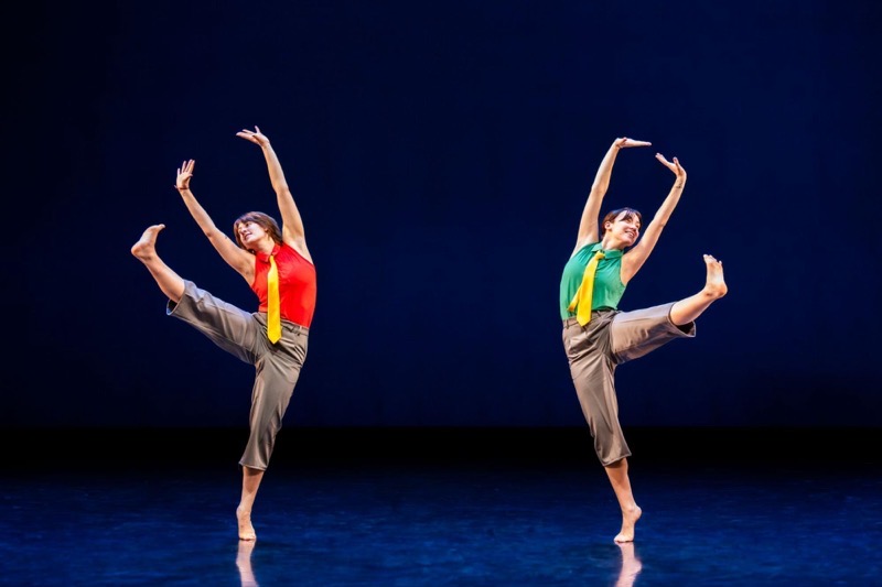 Two figures are depicted in a dynamic dance pose, positioned on a reflective stage. On the left, one figure wears a vibrant red top with a yellow tie, stretching an arm upward while balancing on one leg, with the other leg extended gracefully to the side. On the right, another figure wears a green top and yellow tie, mirroring the pose with a lifted arm and a bent leg, creating a sense of symmetry. The background features a deep blue hue, enhancing the depth of the scene.