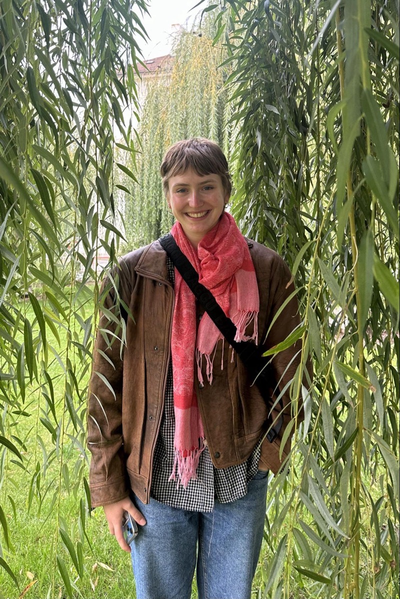 A figure stands smiling amidst long, drooping branches of willow trees, creating a natural frame. The figure wears a brown leather jacket over a black and white checkered shirt, complemented by a bright pink scarf that adds a pop of color. The background reveals a blurred, lush green lawn, enhancing the organic atmosphere of the scene. Soft, diffused lighting suggests an overcast day, contributing to the serene environment.