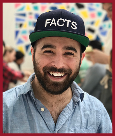 Portrait headshot of a smiling person wearing a dark baseball cap and a light denim shirt, photographed against a softly blurred outdoor background.