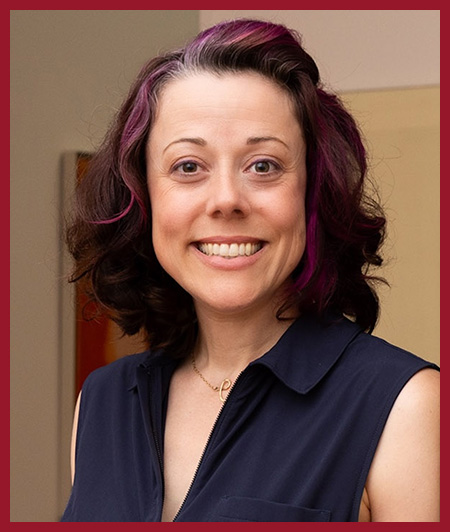 Portrait of a woman smiling at the camera, wearing a dark sleeveless top. She has shoulder-length dark hair and is standing against a muted reddish background.
