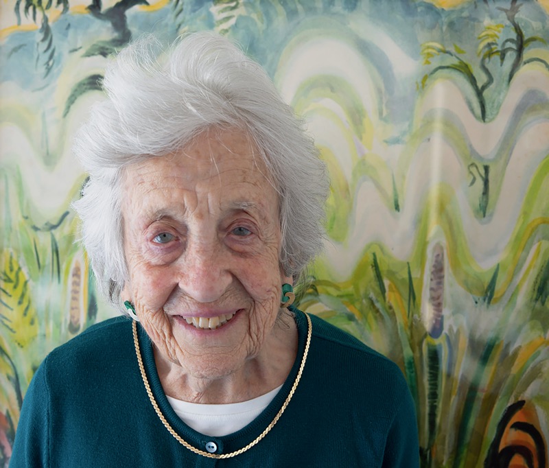 A figure stands in the foreground, smiling warmly, with soft, white hair styled loosely. They are dressed in a dark teal top, accented by a gold chain necklace. Behind the figure, an abstract background features flowing, wavy lines in shades of green, yellow, and hints of brown, resembling organic forms. The lighting is soft and even, creating a gentle atmosphere that complements the vibrant colors of the backdrop.