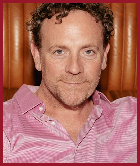 Portrait of a man with short curly hair wearing a pink button-down shirt, smiling slightly at the camera. The background appears to be wood paneling or a warm-toned indoor setting.