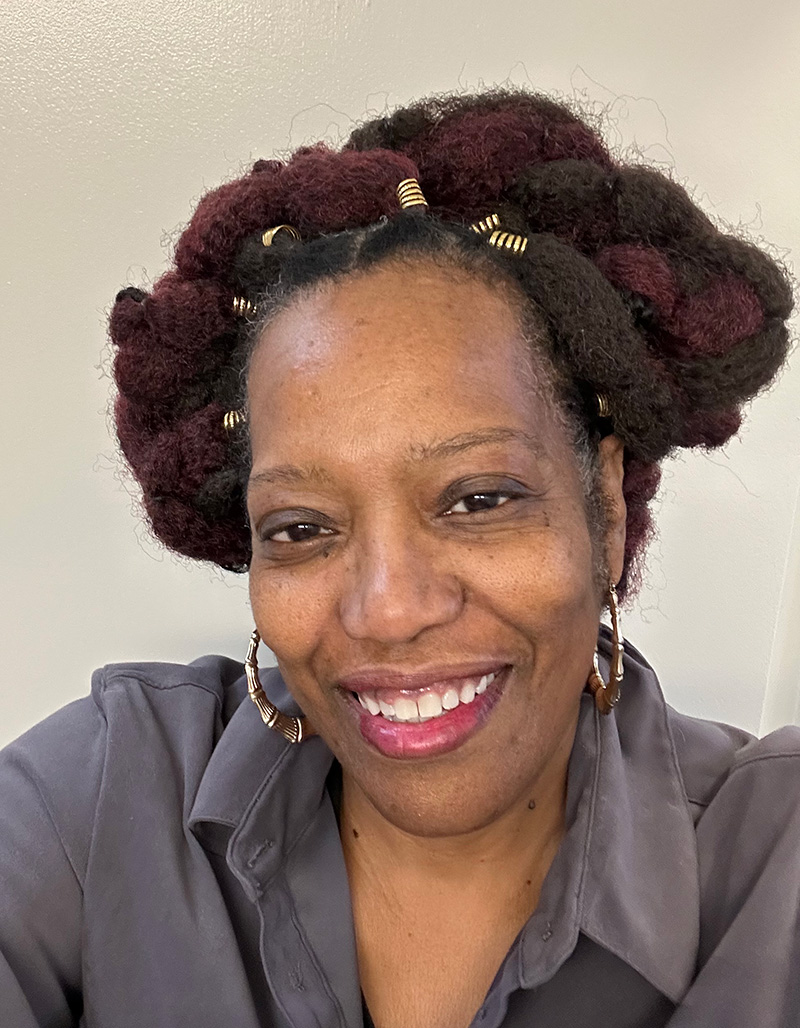 A woman smiling warmly at the camera, wearing a gray button-up shirt and gold hoop earrings. Her hair is styled in burgundy and brown locs pinned up with gold cuffs.