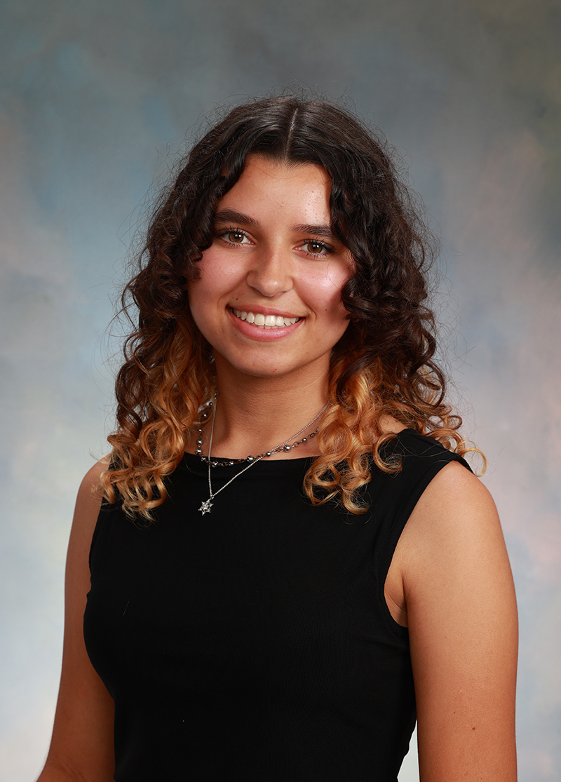 Portrait of a person with shoulder-length curly hair wearing a sleeveless dark top, smiling toward the camera against a neutral studio background.