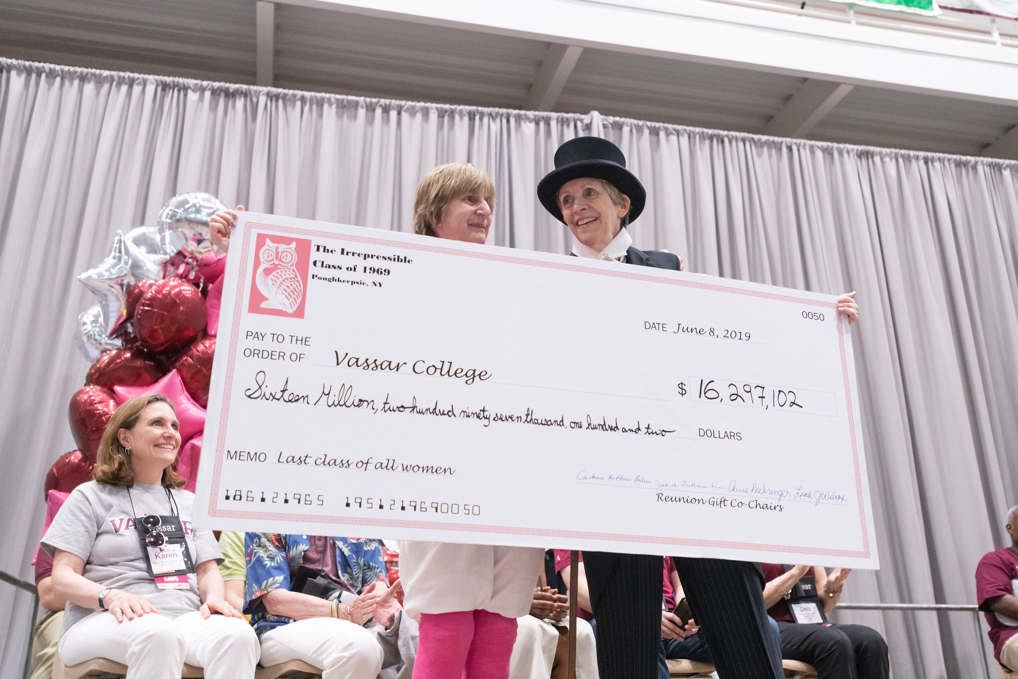 Two alums with a large check for 16 million, one dressed in a top hat and tux..