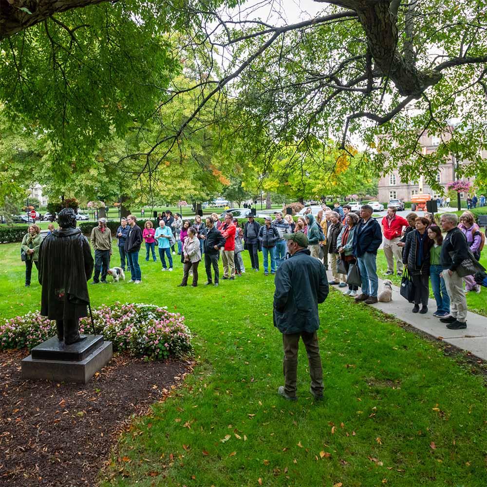 A group gathers under shade trees on a campus lawn, facing toward a bronze statue on a pedestal in the foreground, listening to a speaker facing them..