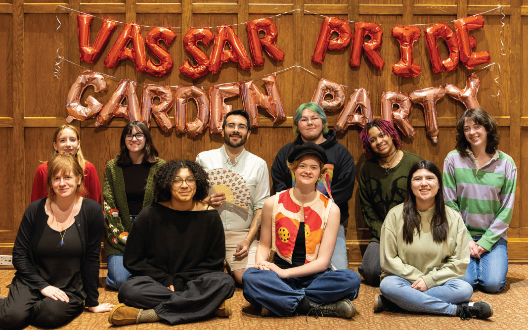 Group photo of nine people smiling under "Vassar Pride Garden Party" balloons against a wood wall.