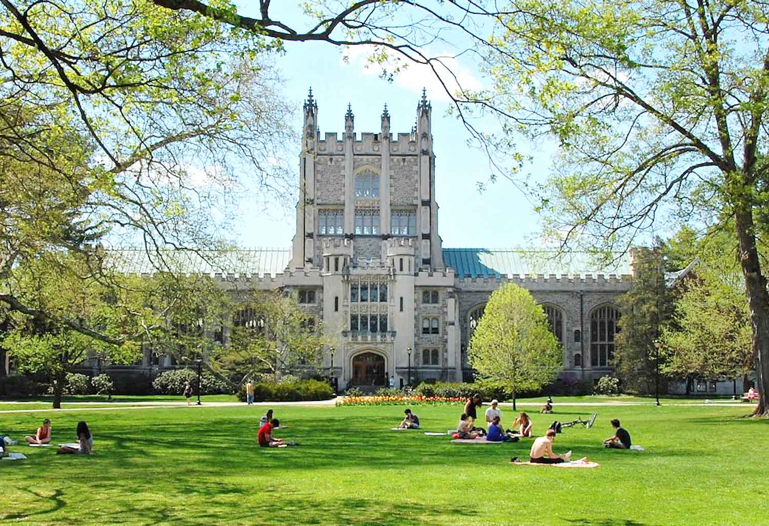 Seated groups of people scattered on a green lawn in front of the library building on a college campus.