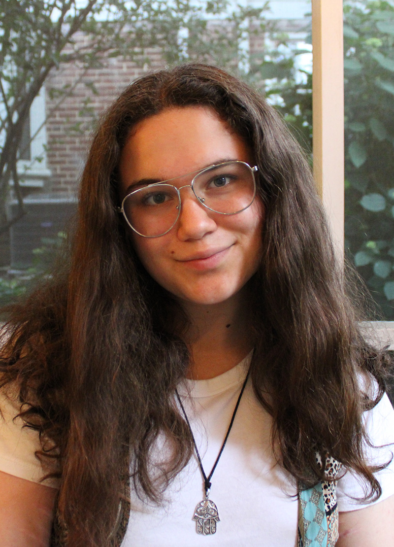 Person with long dark hair and glasses, wearing a white shirt and pendant necklace, seated indoors in front of a window with trees visible outside.