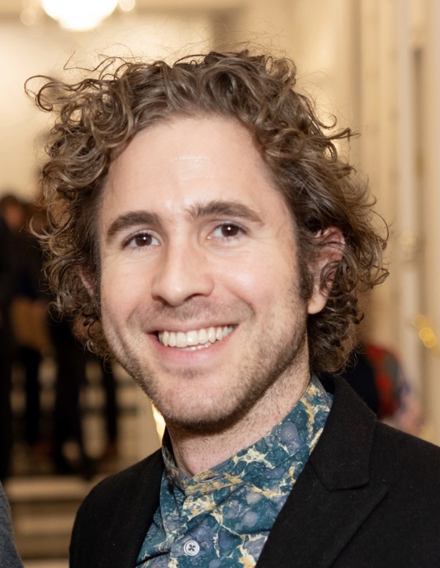A portrait of Patrick Gallagher, a person with curly hair, wearing a dark blazer over a brightly patterned shirt with shades of blue and green, resembling abstract marbling. The individual is smiling, presenting an inviting expression. The background is softly blurred, with hints of warm lighting and indistinct figures, suggesting a gathering or event. The overall tone is light and social, with a focus on the subject in the foreground.