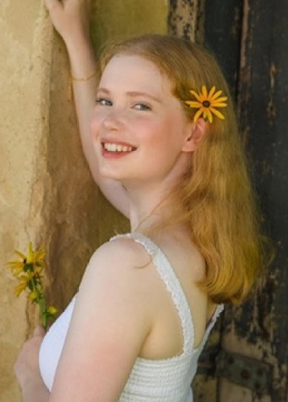 Person with shoulder-length light hair wearing a sleeveless white top, smiling toward the camera while standing beside a stone wall, with yellow flowers tucked into their hair and held in one hand.