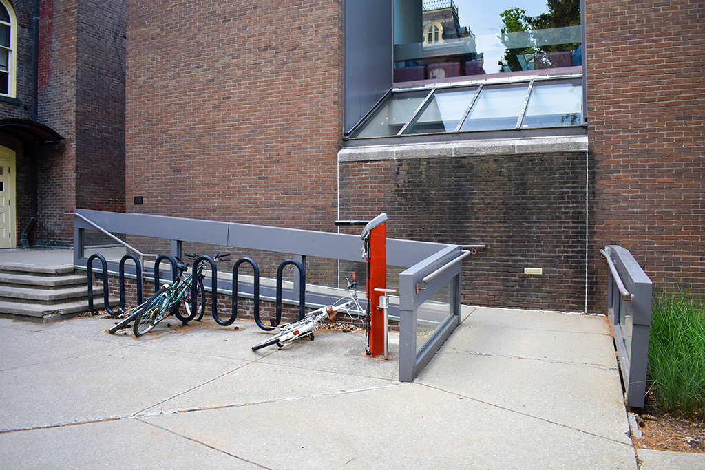 Exterior view of a low-grade ramp to the east of a few stairs, with a bike rack in between.
