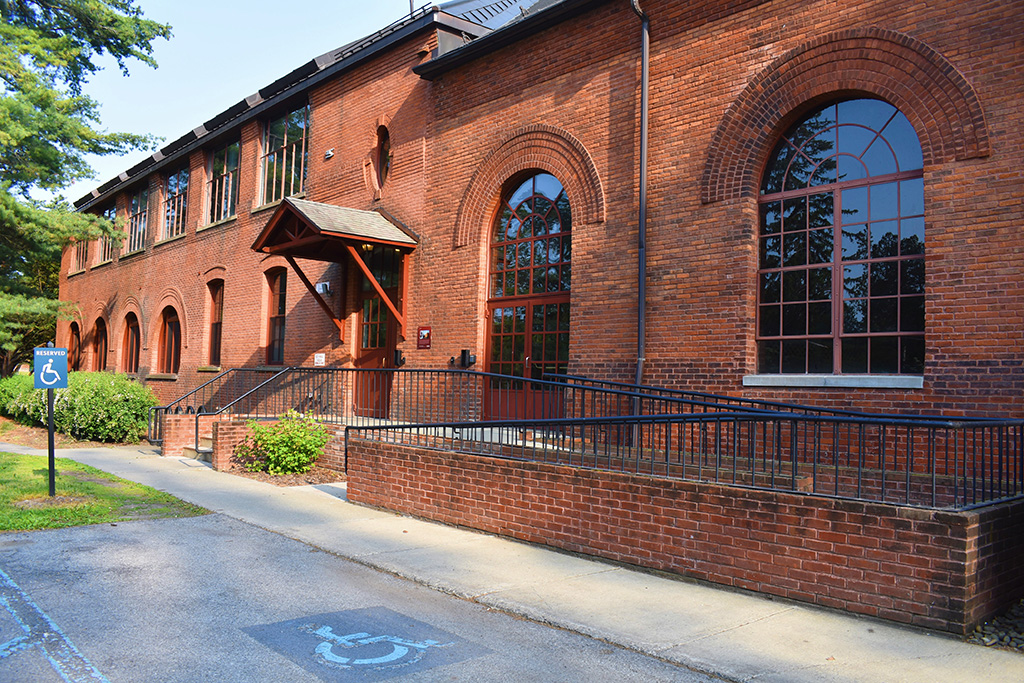 An exterior shot of a large brick building with arched windows and a central entrance with a small canopy and a ramp leading to it.