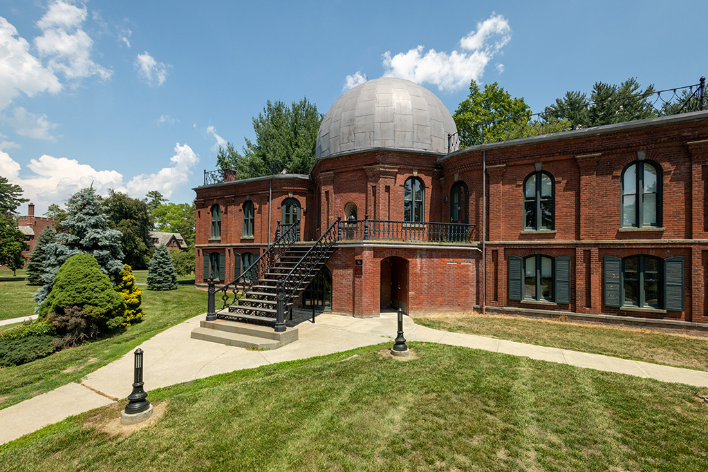 Exterior view of a historic red-brick observatory building with arched windows, a central staircase leading to a domed roof, and surrounding green lawns under a bright blue sky with scattered clouds.