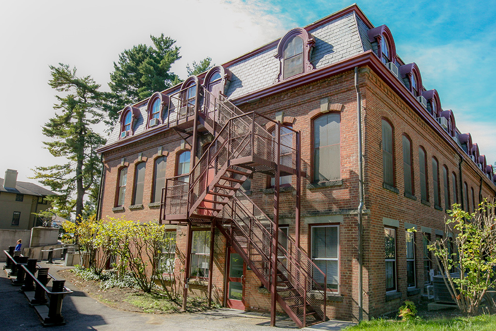 Historic red-brick building with mansard roof and decorative dormer windows, featuring an exterior metal staircase on the corner and surrounded by landscaping under a bright blue sky.