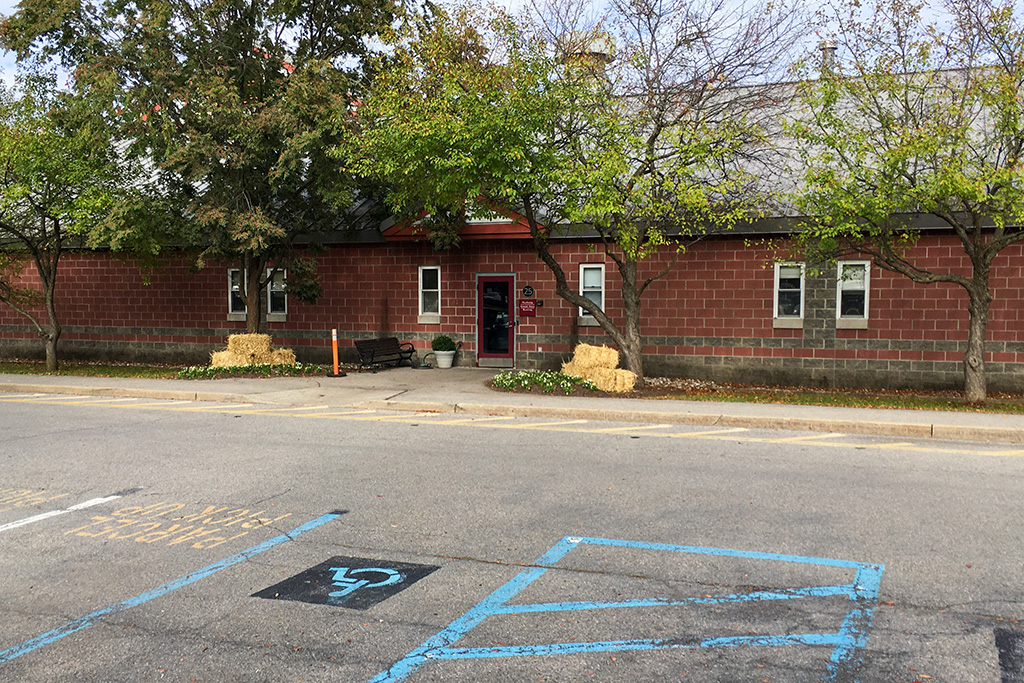 Single-story red brick building with a gray roof, small rectangular windows, and a central entrance door. The front is decorated with potted plants and trees partially shading the facade. An accessible parking space is painted in blue in the foreground.
