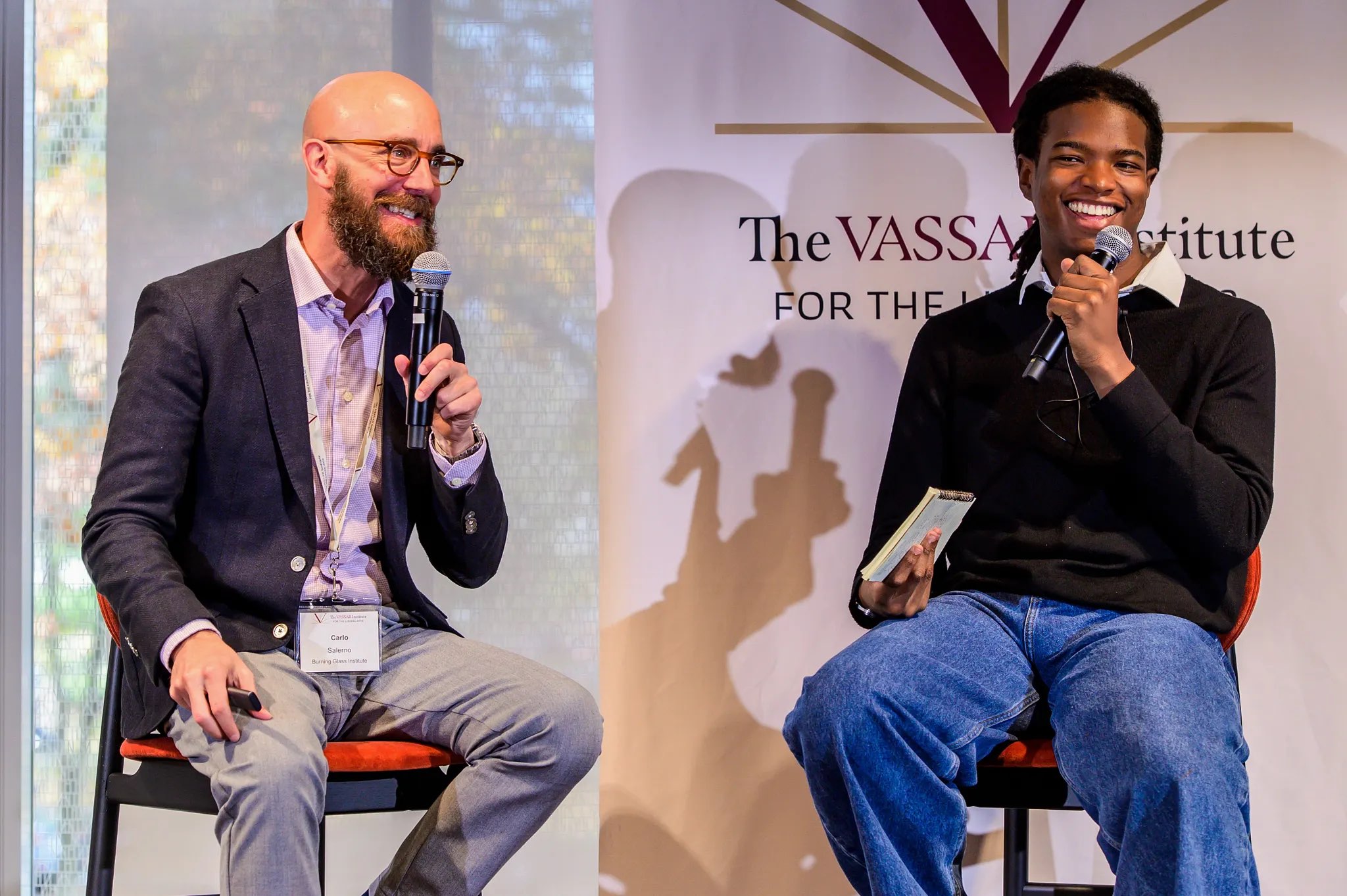 Two individuals are seated on stage, engaged in a conversation. One is holding a microphone and smiling, while the other is speaking into a microphone and gesturing with their hands. The background features a decorative banner with the text "The Vassar Institute for the Arts." Natural light streams in from large windows, creating a bright atmosphere, while the stage setup includes simple chairs and a minimalistic decor.