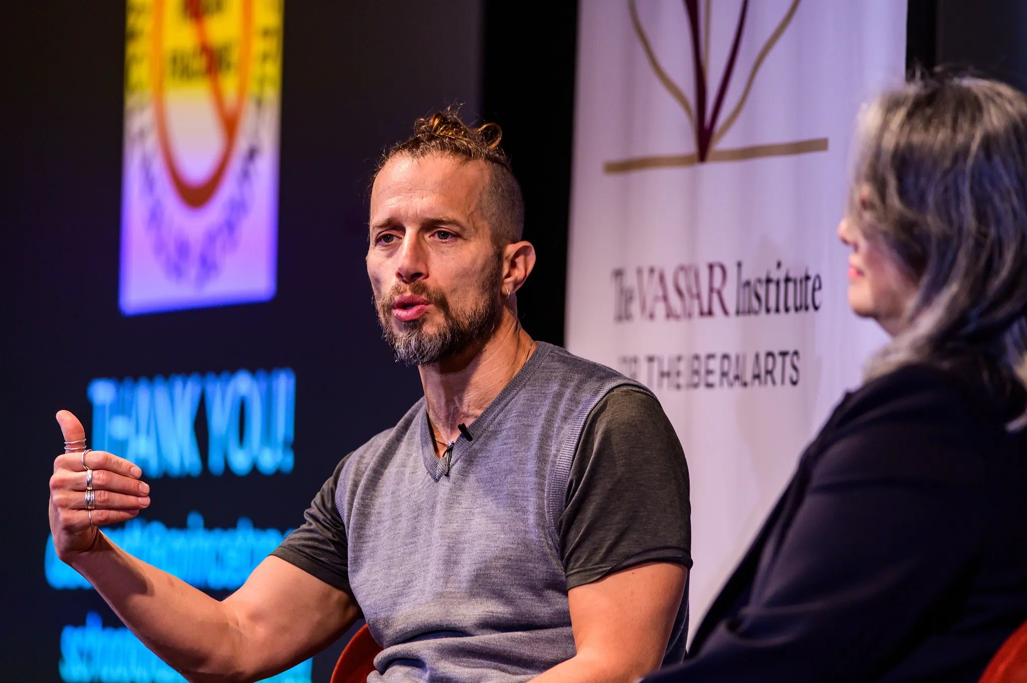 A person with a beard and short hair is seated, gesturing with their left hand while speaking. They are dressed in a gray shirt and exhibit a focused expression. To their right, another individual listens attentively, partially visible, with lighter hair styled down. The background features a digital screen displaying text in blue and an emblem above, along with a presentation backdrop that includes the Vassar Institute logo in muted colors. The lighting is bright, illuminating the speakers clearly.