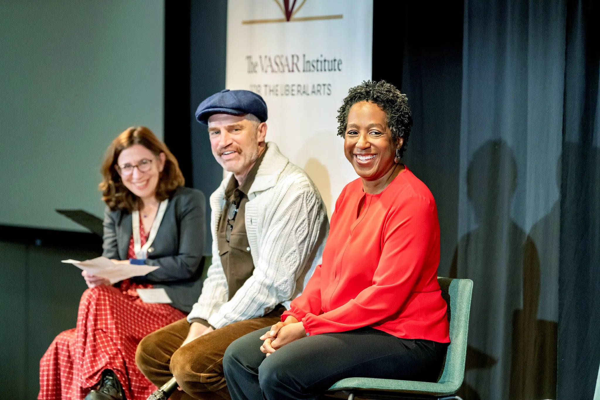 A panel discussion takes place on a stage. Three individuals are seated on chairs, with two on the left and one on the right. The left figure wears a patterned red dress and glasses, holding papers and smiling. The central figure is dressed in a light jacket and hat, while the figure on the right wears a bright red top. The backdrop features a banner with the text "The Vassar Institute for the Liberal Arts." Soft lighting highlights the subjects against a dark background.