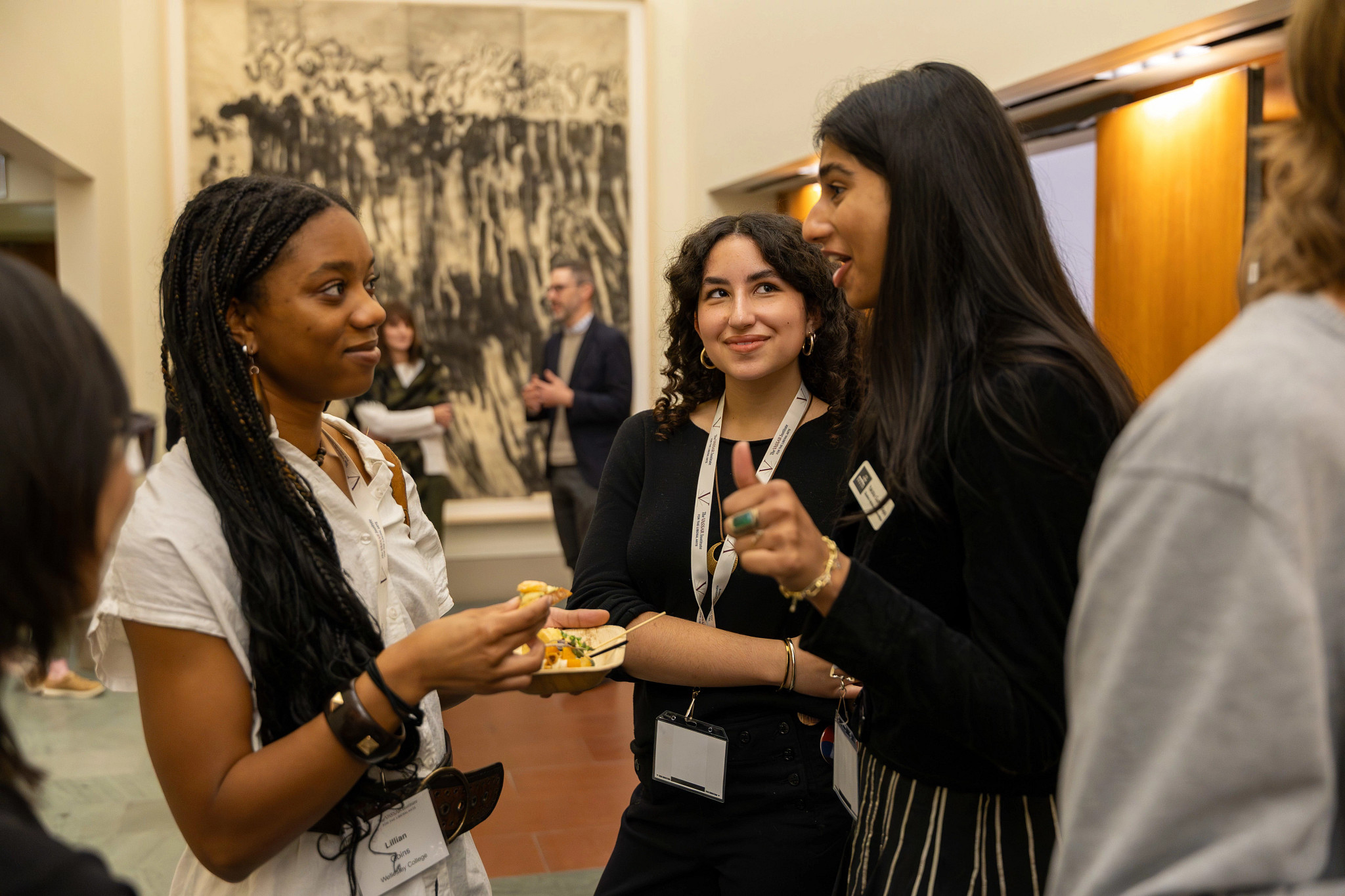 A group of three individuals engage in conversation in a well-lit indoor space. One figure, positioned on the left, holds a plate of food and looks attentively at another, who is speaking animatedly. The third person, standing near them, smiles while listening. The background features a large abstract artwork in muted tones, enhancing the vibrant colors of the participants' clothing, with wooden surfaces and contemporary decor visible throughout the setting.