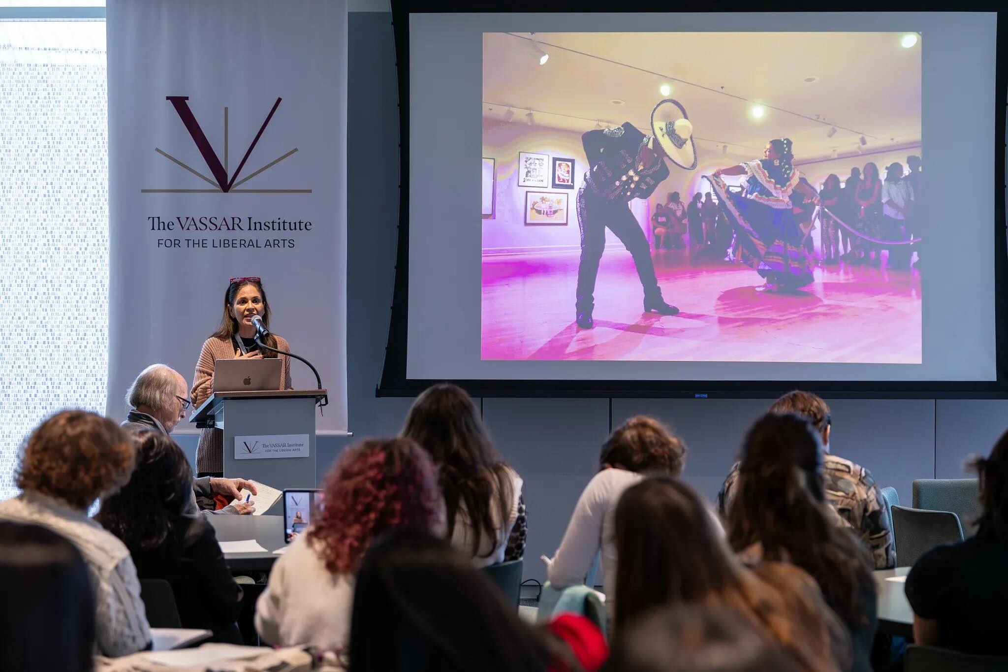 In a spacious auditorium, a speaker stands at a podium in front of a large screen displaying dancers in traditional attire, performing on a wooden floor illuminated by soft purple lighting. The speaker, dressed in neutral tones, addresses an audience seated at tables, which are cluttered with papers and materials. The audience consists of various figures, focused on the presentation.