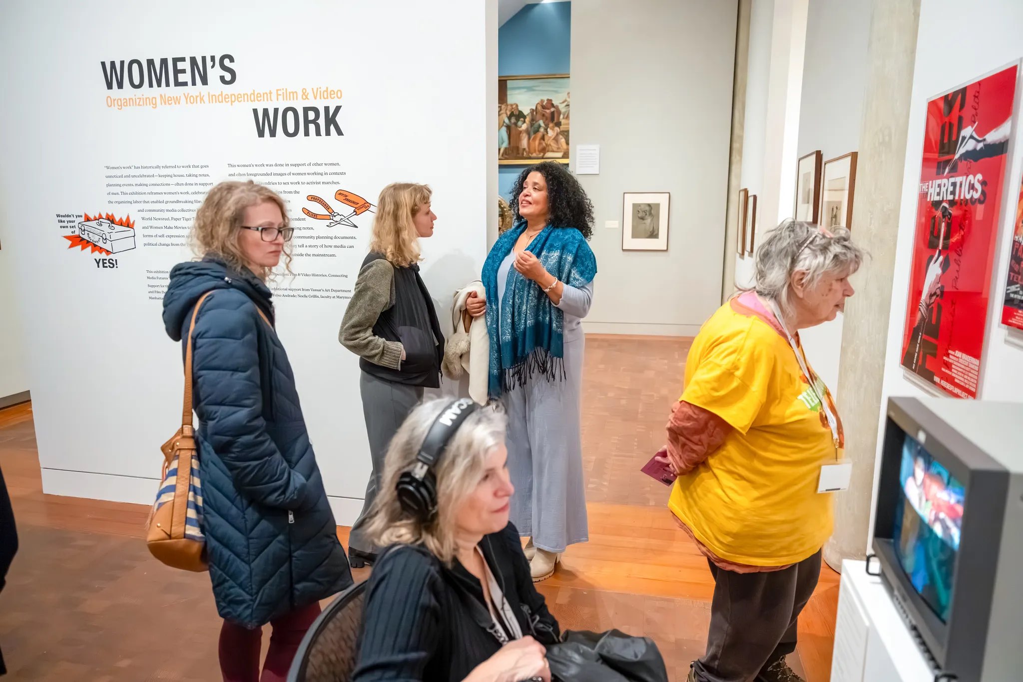 In a gallery setting, several figures are engaged with an exhibit titled "Women's Work." The wall display features text and images related to independent film and video. One individual in a blue shawl interacts animatedly, while another stands nearby, wearing a green top. To the left, a figure with headphones listens intently, and another in a yellow shirt stands near a framed poster titled "The Heretics," alongside a vintage television showing video content.