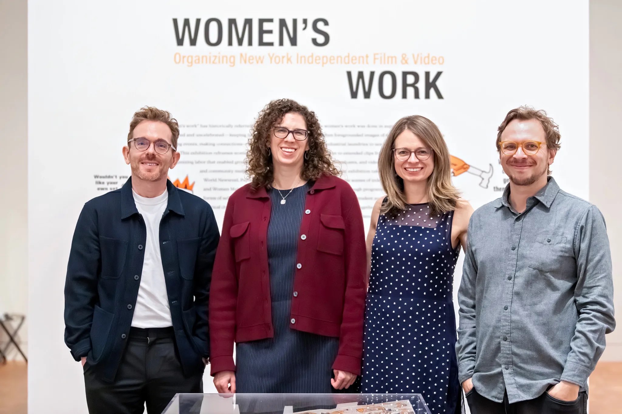 Four individuals stand in front of a large wall with the text “Women’s Work” prominently displayed at the top, with "Organizing New York Independent Film & Video." The figures wear casual attire, with varied accessories including eyeglasses and a mix of colors in their clothing. A glass display case in the foreground contains items related to the exhibit, while soft, even lighting enhances the overall atmosphere of the setting.