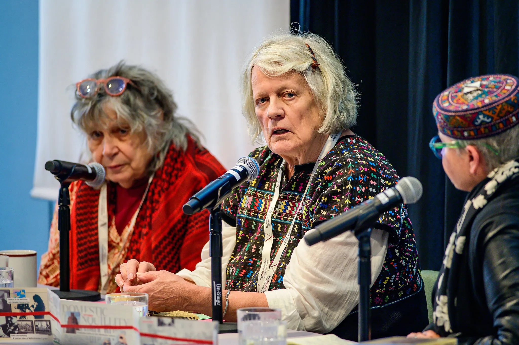 A panel discussion is underway, featuring three figures seated at a table with microphones. One figure in the center, wearing a colorful patterned garment, is speaking, while the others listen attentively. The setting is well-lit, with a soft focus background that suggests an indoor space with muted colors.