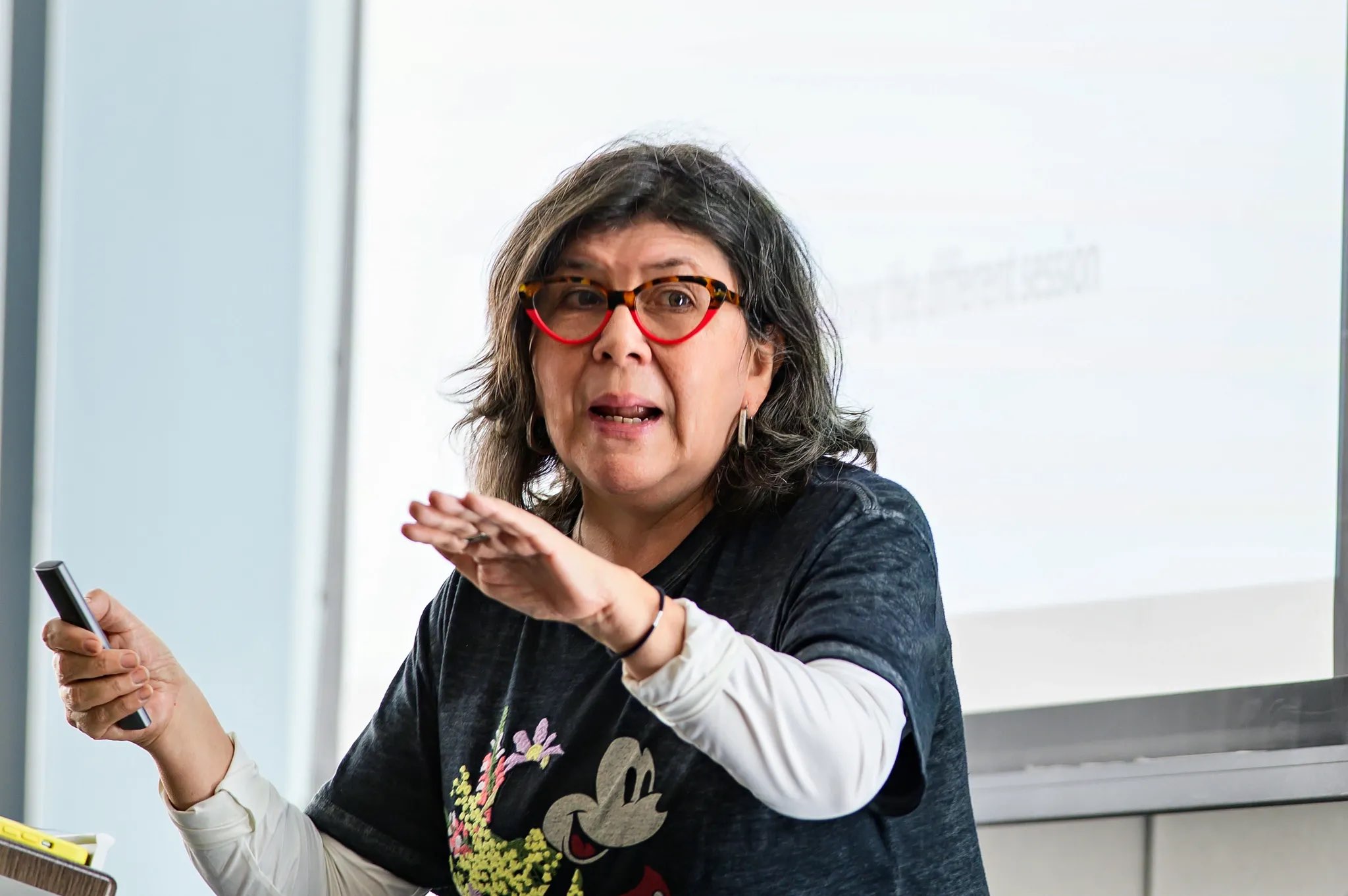 A person stands in foreground, engaging with an audience while gesturing with one hand. They are wearing a dark t-shirt featuring a graphic of a character and white long sleeves underneath. The setting is bright, with natural light coming from large windows in the background, creating a light atmosphere. A presentation screen is visible behind them, displaying text that is not legible.