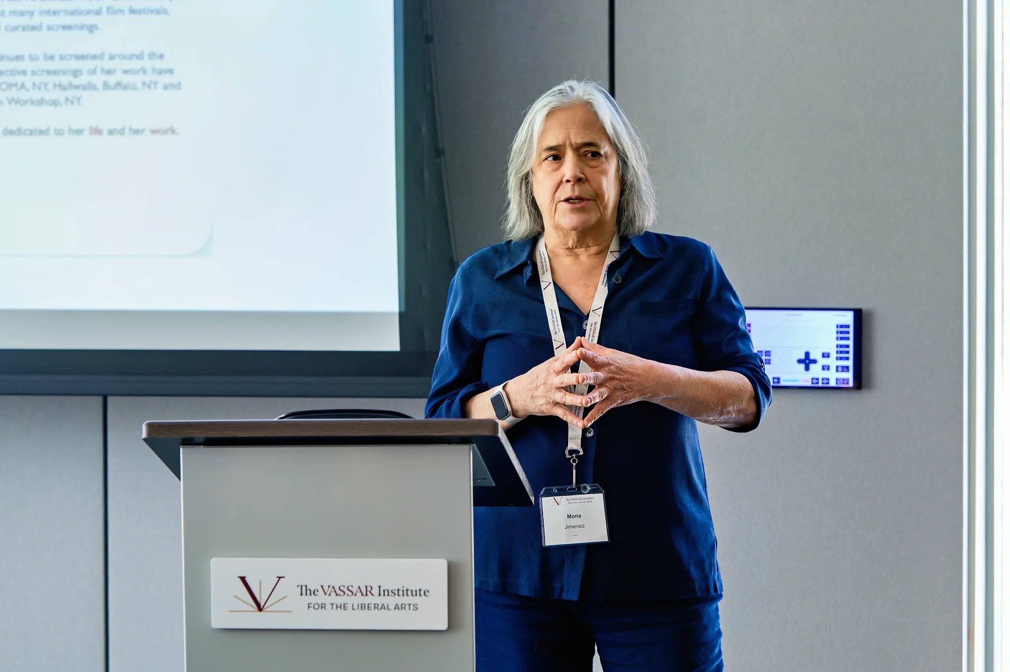 A figure stands in front of a presentation screen, delivering a talk at a podium marked with the logo of The Vassar Institute for the Liberal Arts. The figure is dressed in a dark blue outfit with a name tag visible, gesturing expressively with hands. The background features a sleek, modern environment with neutral-colored walls and a digital display. 