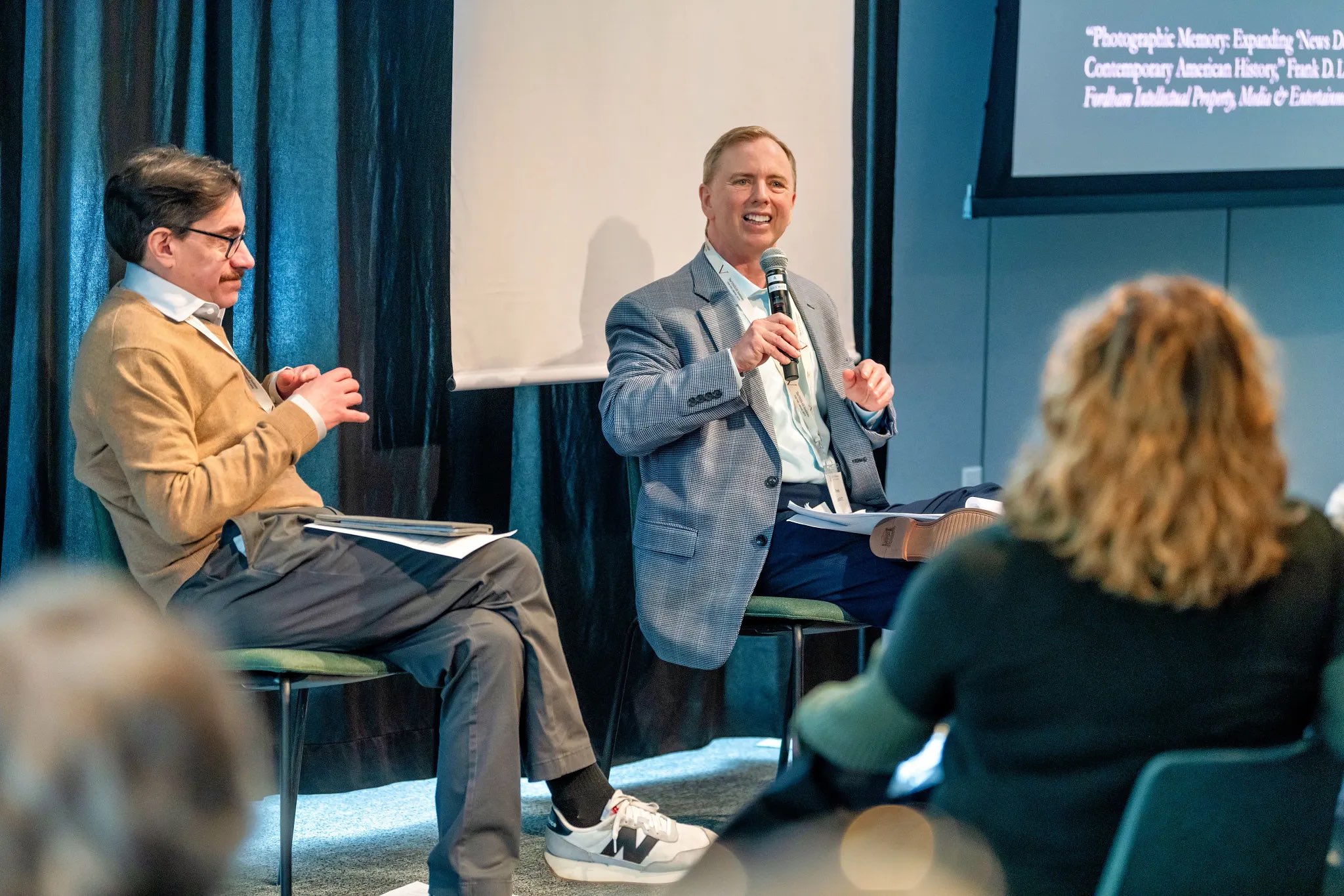 Two individuals are seated in a modern indoor setting, engaging in a conversation. One person, wearing a light gray blazer over a blue shirt, is smiling and holding a microphone. The other individual, in a beige sweater, listens with a thoughtful expression, seated with hands resting on their knees. The background features dark curtains, and a screen displays text related to the discussion topic, creating a professional atmosphere with soft, focused lighting.