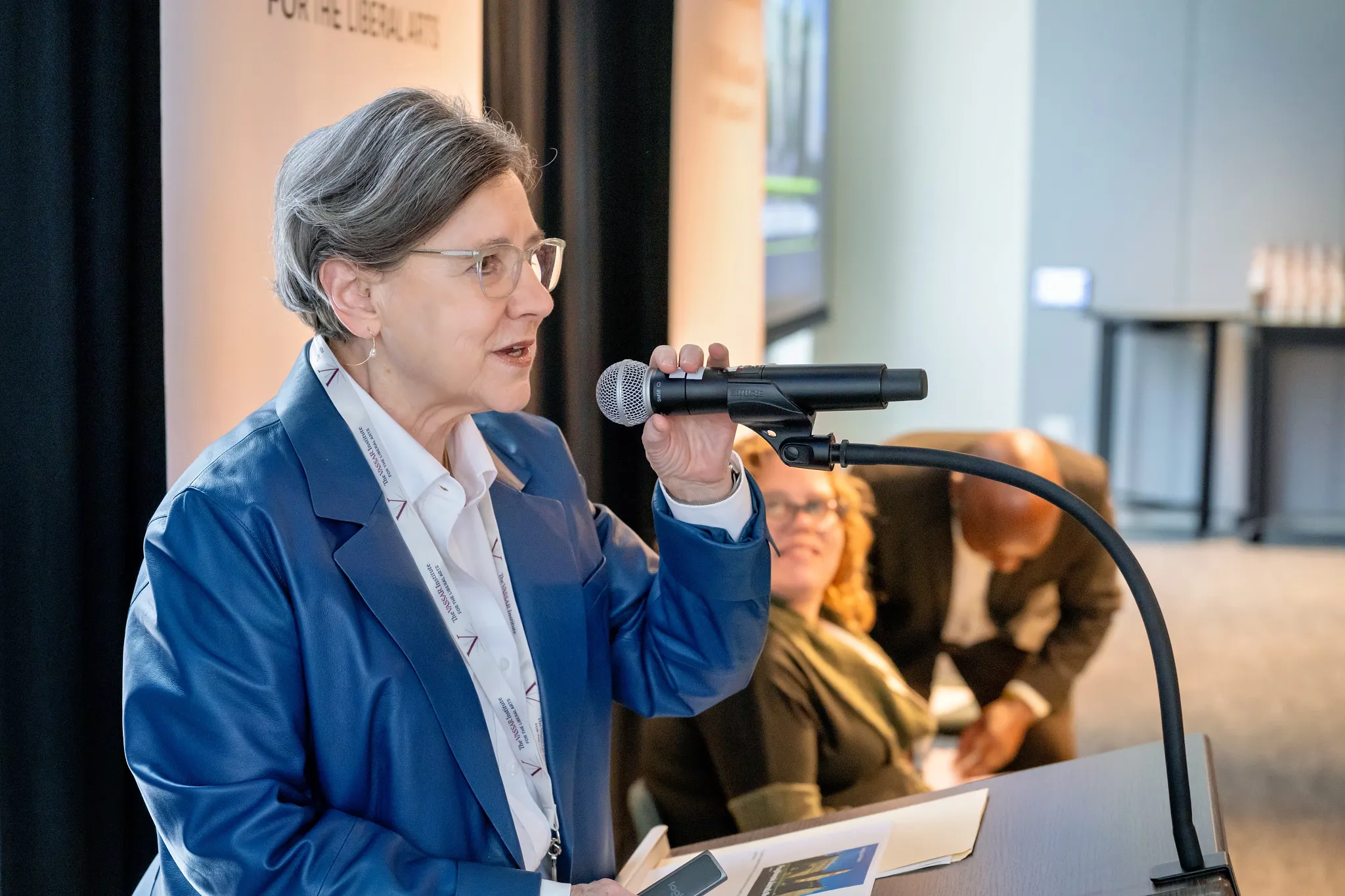 A person in a blue blazer and glasses speaks into a microphone at a podium during an event.