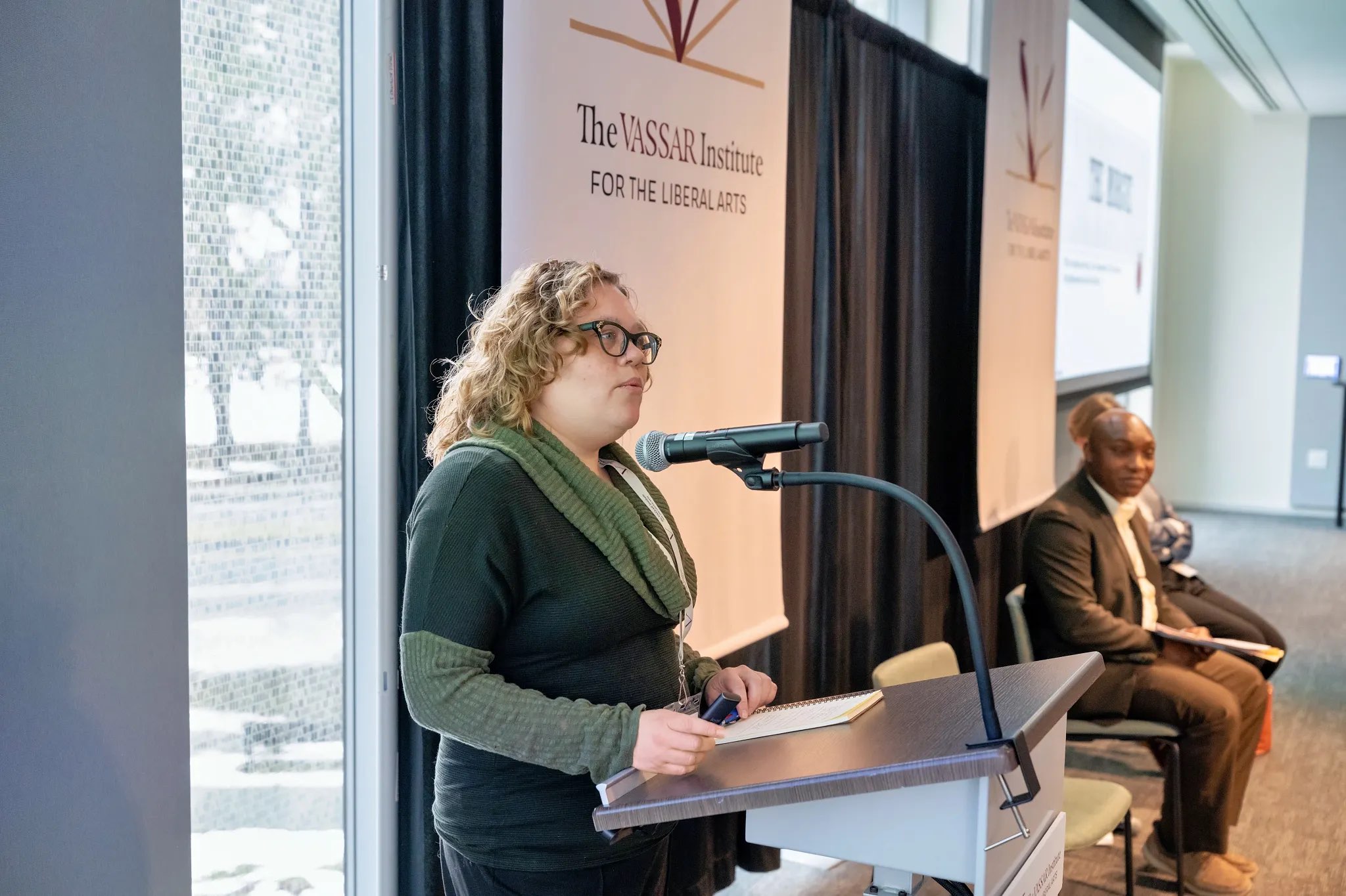 A speaker stands at a podium equipped with a microphone, facing an audience. The individual wears glasses and a dark green sweater, holding notes in one hand while gesturing slightly with the other. Behind the speaker, there are two large banners displaying the logo of the Vassar Institute for the Liberal Arts. In the background, another person sits in a chair, while natural light filters in through large windows, creating a soft ambiance in the room.