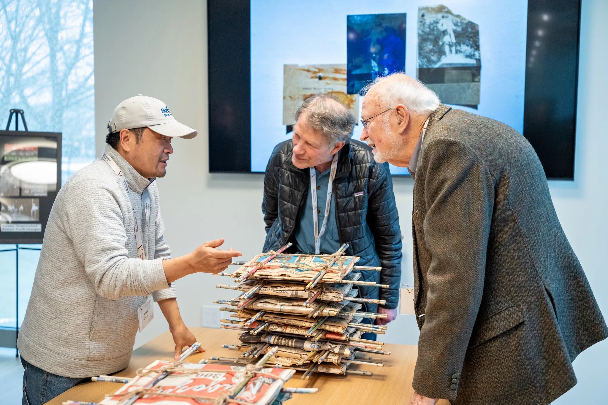 A group of three individuals is gathered around a wooden table displaying a stack of flat, layered objects secured with sticks. One person, wearing a gray sweater and cap, gestures while speaking, engaging the others. The second individual, dressed in a black padded jacket, leans closer, appearing intrigued, while the third, in a brown tweed jacket, observes intently. In the background, two framed images with varying colors and textures are mounted on the wall.