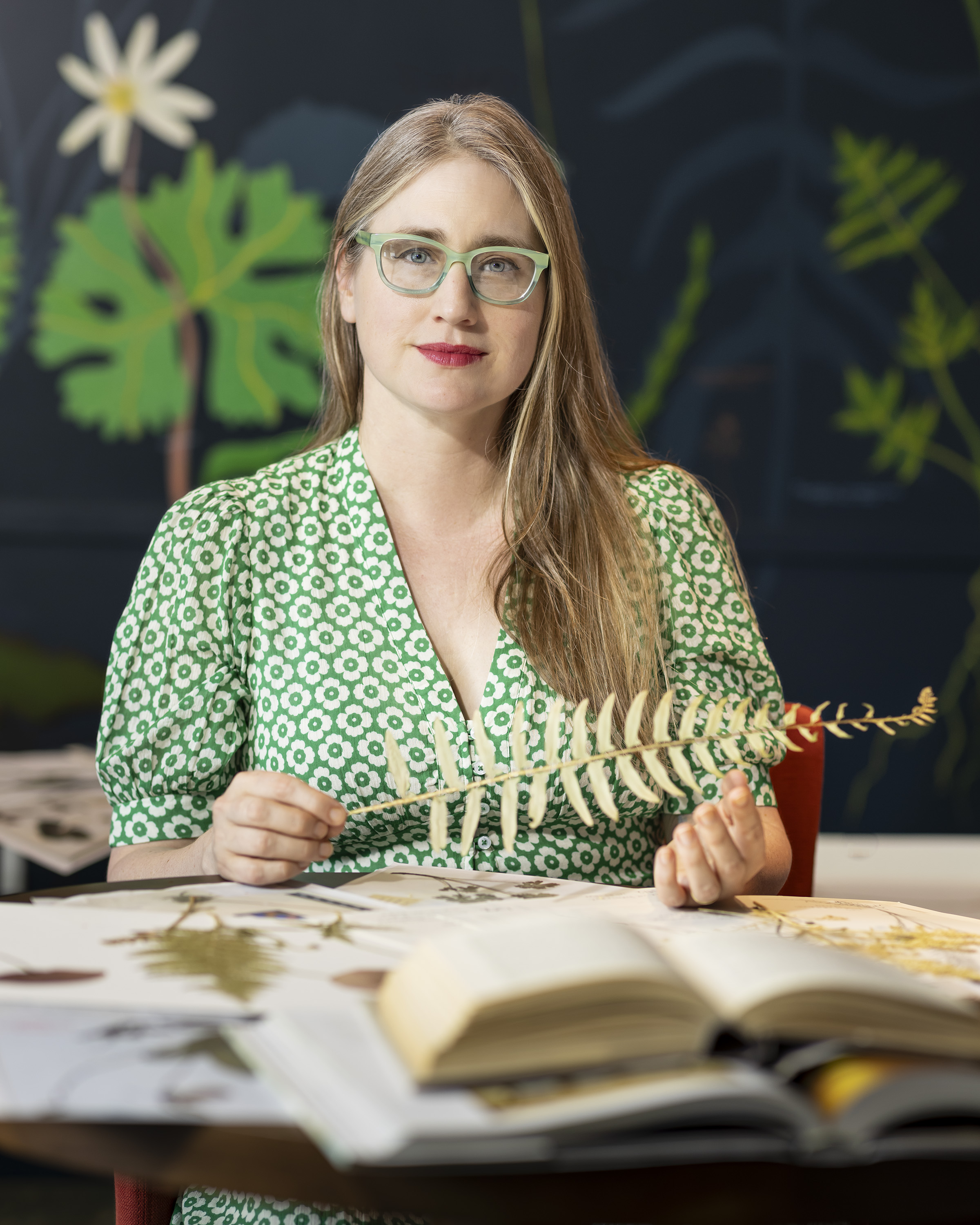 A person sitting at a table with botanical books and holding a dried fern. They look out at the viewer. 