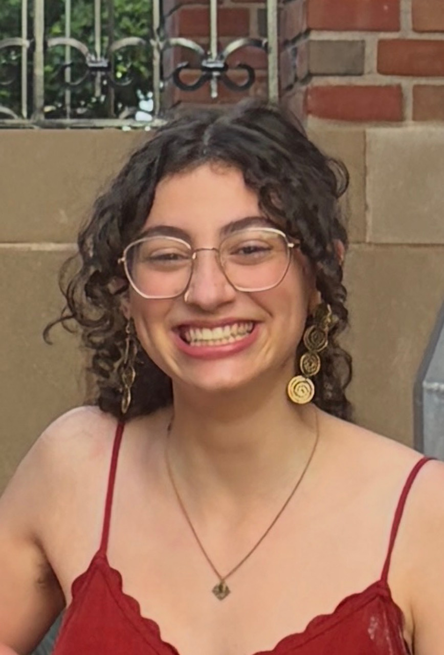 Em Friedman with shoulder-length curly hair, thin-framed glasses, and gold dangle earrings, wearing a red spaghetti-strap top outdoors against a stone and brick wall background.
