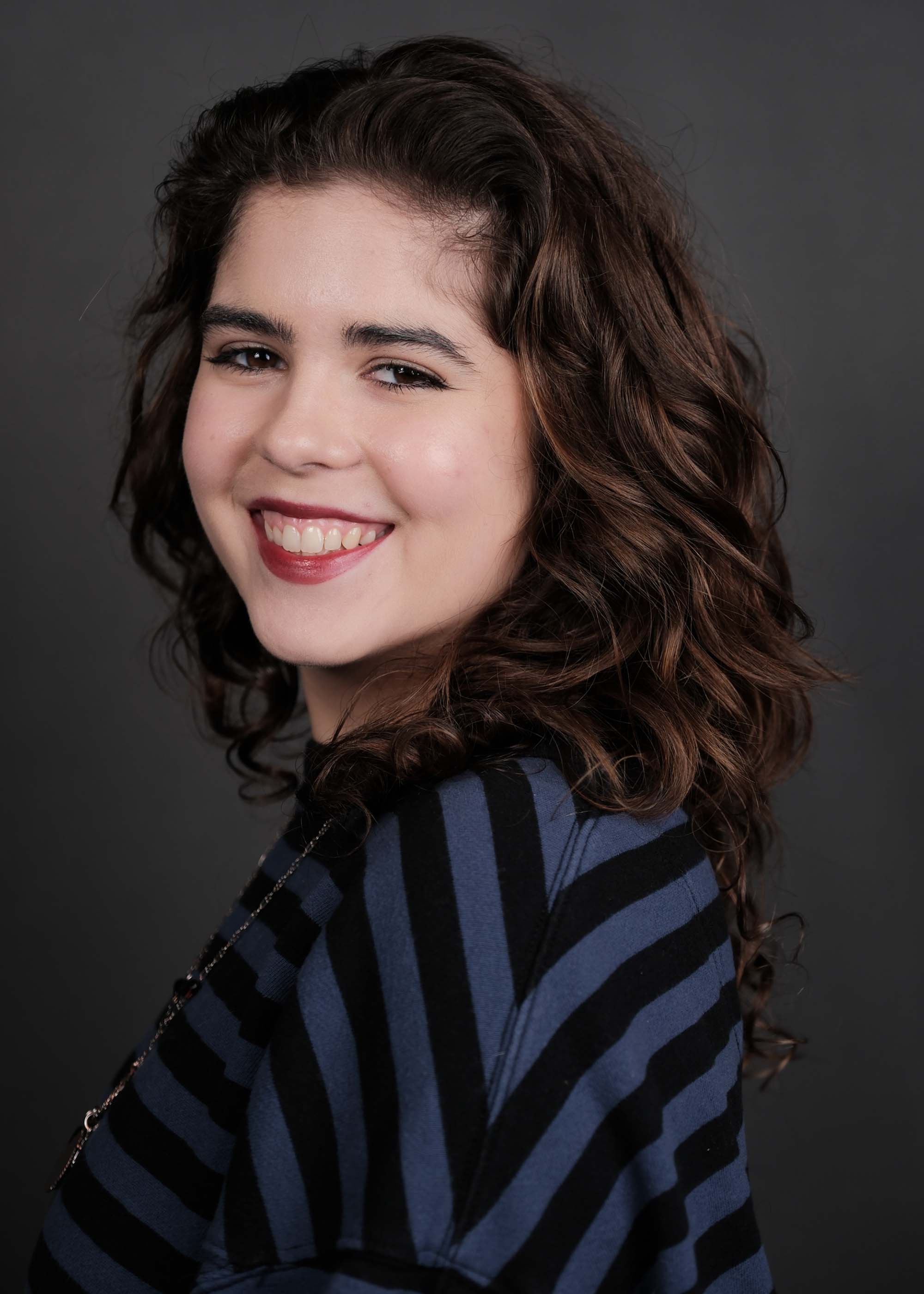 Mia Zottoli with curly brown hair looking over her shoulder with a broad smile, wearing a dark blue and black diagonally striped top set against a solid dark gray backdrop.