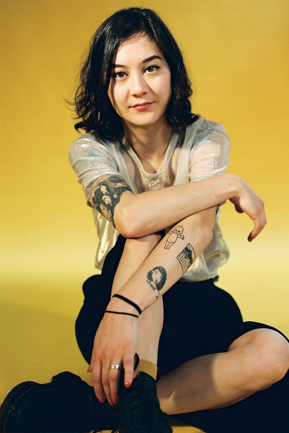 Michelle Zauner seated against a yellow background, wearing a sheer top and black skirt, with several tattoos visible on their arms.