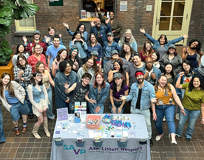 A large group poses enthusiastically behind an information table with Support, Advocacy, & Violence Prevention Office materials and a banner reading "SAVP Ask. Listen. Respect."