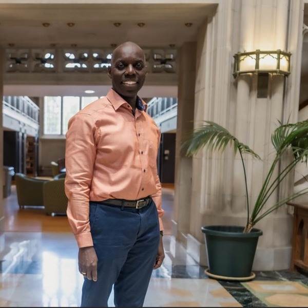 A person in a salmon button-down shirt and dark trousers stands in a bright interior hallway. Tall columns, patterned stone flooring, wall lamps, and potted palms frame the figure. Warm indoor light mixes with daylight entering through windows and glass doors.