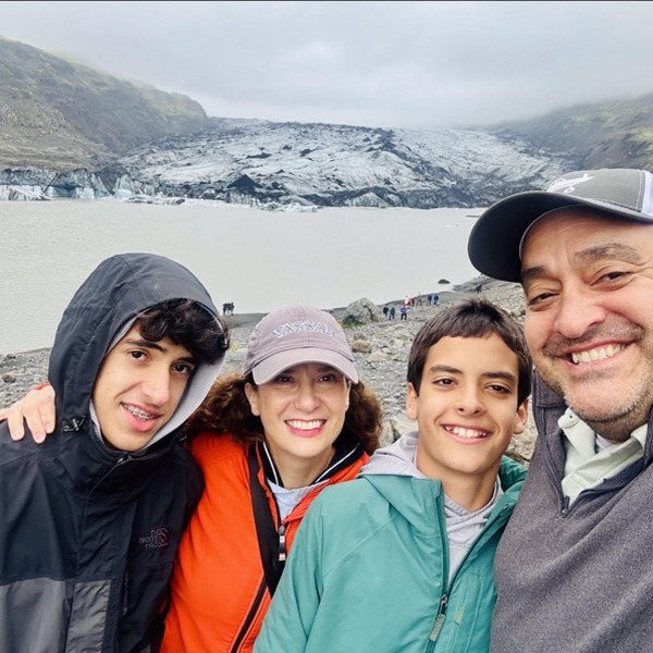 Four people stand closely together at a glacial overlook, with arms resting around one another. A pale lake, dark rocky slopes, and a broad glacier stretch across the background. Gray clouds diffuse the light over the mountainous landscape.